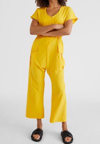 Gul jumpsuit med korta ärmar, rundad halsringning och elastisk midja. Har två sidofickor och bred bendesign. Bärs med svarta sandaler med öppen tå.