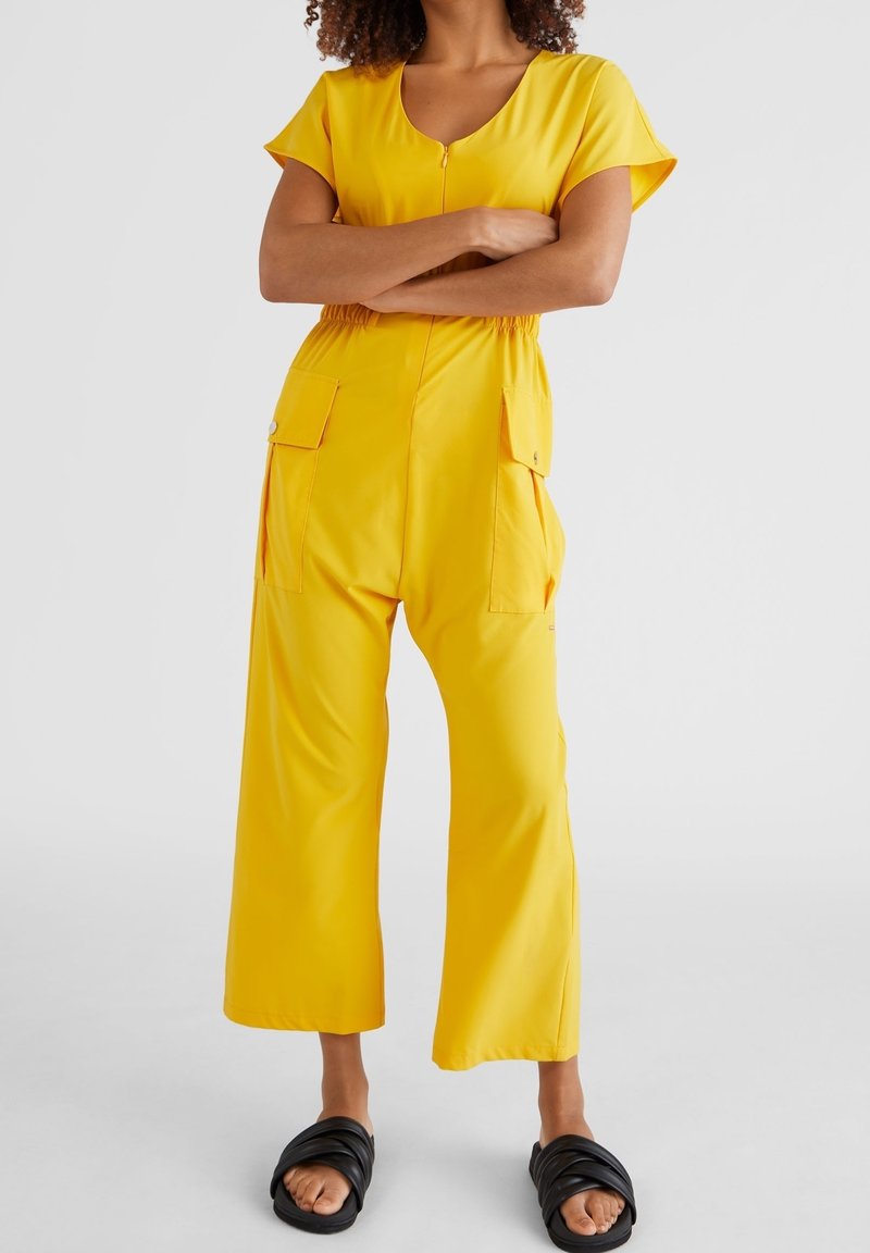 Gul jumpsuit med korta ärmar, rundad halsringning och elastisk midja. Har två sidofickor och bred bendesign. Bärs med svarta sandaler med öppen tå.