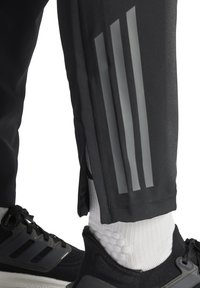 Schwarze Sporthose mit grauem reflektierendem Drei-Streifen-Detail, mit einem Reißverschluss an der Seite, kombiniert mit schwarzen Turnschuhen und weißen Socken.