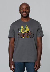 Grijze t-shirt met een cartoonontwerp van twee avocado's die op een rode tandemfiets rijden, met gele accenten en zwarte omlijningen.