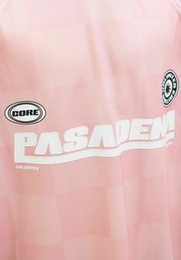 Jersey deportivo rosa claro con textura a cuadros, "PASADENA" en letras blancas y el logo "CORE" arriba, con un detalle de insignia redonda.