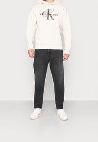 Sudadera con capucha de un beige claro con "Calvin Klein Jeans" impreso en gris oscuro, combinada con jeans negros y zapatillas blancas. Ajuste casual.