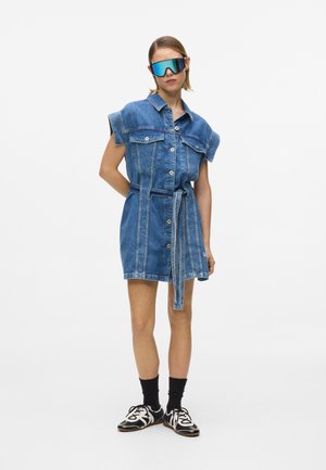 Donna che indossa un vestito di jeans senza maniche con cintura, calzini neri, sneaker bianche e nere e occhiali da sole oversize riflettenti, in piedi contro uno sfondo bianco.