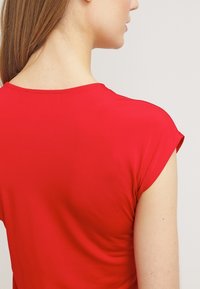 Haut rouge à manches courtes en tissu lisse, avec un col rond et un ourlet légèrement arrondi à l'arrière. Aucun motif ou accent visible.