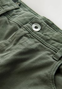 Close-up roheline denim pükste detail, kus on näha nööp, millel on reljeefne tekst "DENIM SUPPLY Co." ja nähtav õmblus vöökoha ja vöörõnga lähedal.