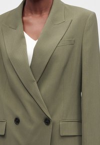 Gros plan d'une personne portant un blazer croisé à fines rayures vert olive clair avec des boutons noirs et un haut blanc en dessous.