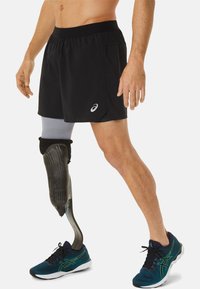 Schwarze Sportshorts mit einem elastischen Bund und einem grauen Prothesenbein. Die Person trägt blaue Turnschuhe mit grünen Akzenten.