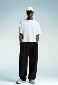 Hvid bomulds T-shirt, brede sorte corduroybukser, grå sneakers og en hvid cap med maroon accenter. En simpel, afslappet pasform og materialer.
