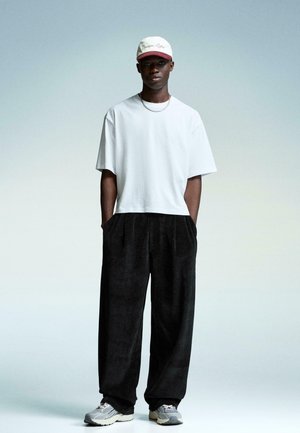 White cotton T-shirt, wide black corduroy trousers, gray sneakers, and a white cap with maroon accents. Simple, relaxed fit and materials.