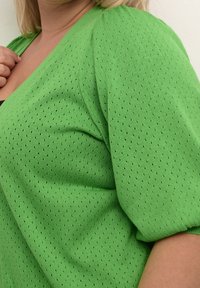 Cardigan en tricot vert avec des manches bouffantes courtes, présentant une texture perforée et un décolleté en V. Des accents noirs sont visibles dans les coutures.