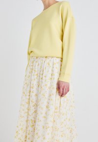 Geel sweatshirt met lange mouwen, gecombineerd met een witte plisseerrok met gele bloemmotieven. Zachte textuur en casual pasvorm.