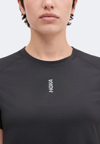 Κοντινό πλάνο ενός ατόμου που φοράει μαύρο t-shirt Hoka, δείχνοντας τον λαιμό, το ανώτερο μέρος του στήθους και το λογότυπο σε μονόχρωμο φόντο.