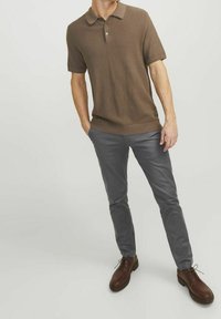 Camisa polo marrón de manga corta con un tejido texturizado y cierre de tres botones, combinada con pantalones gris ajustados y zapatos de cuero marrones con cordones.