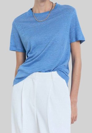 Woman wearing a light blue short-sleeve shirt tucked into high-waisted white pants with a silver chain necklace.