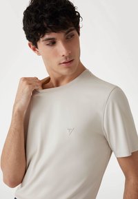 Hellbeige T-Shirt mit kurzen Ärmeln aus glattem Stoff, ausgestattet mit einem kleinen dreieckigen Logo auf der linken Brust und einem Rundhalsausschnitt.