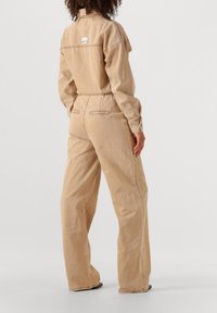 Persoon met krullend haar draagt een losse beige denim overall, staat met de rug naar de camera gericht, op een effen witte achtergrond.