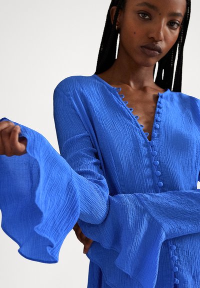 Γυναίκα με πλεγμένα μαλλιά που φοράει ένα φωτεινό μπλε φόρεμα με υφή, κουμπιά στο μπροστινό μέρος και φαρδιά, μακριά μανίκια με βολάν.