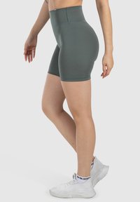 Pantalones cortos verdes de talle alto y ajustados, hechos de material elástico con una textura suave y costuras limpias. Llevados con zapatillas deportivas blancas.