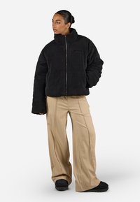Giacca in pile nera con colletto alto, zip frontale e maniche larghe. Indossata con pantaloni beige a gamba larga e calzature nere. Sfondo semplice.