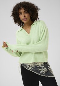 Pull vert clair avec col en V et col, manches longues et coupe décontractée, superposé à un vêtement noir orné de dentelle.