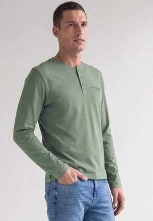 Homme portant une chemise henley verte à manches longues et un jean bleu, debout avec une main dans la poche sur un fond uni.