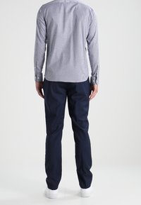 Mężczyzna stojący tyłem, ubrany w jasnoniebieską koszulę w kratkę z długim rękawem, granatowe spodnie i białe sneakersy na jednolitym tle.