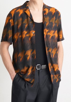 Man draagt een zwart-oranje patroon knoopshirt met korte mouwen over een zwart singletje, met zwarte broek en zwarte riem.