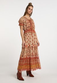 Pitkä, lennokas mekko beige värissä, jossa on punaisia kukka- ja paisley-kuvioita. Ominaisuuksiin kuuluu kerätty pääntie, lyhyet hihat ja kerrostettu hame.