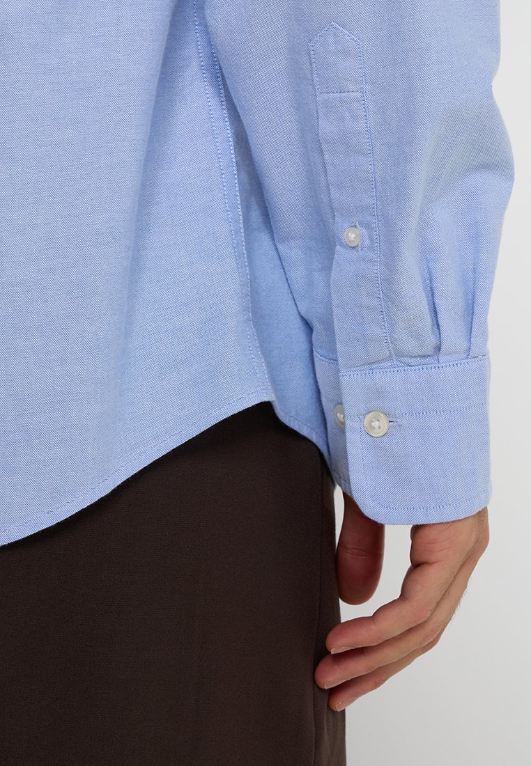 Jasnoniebieska koszula z długim rękawem, z zapięciem na guziki przy mankietach, uszyta z teksturowanej tkaniny, z bocznymi rozcięciami i zaokrąglonym dołem.