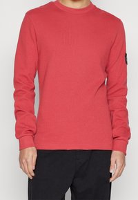 Röd långärmad skjorta med en strukturerad ribbad design, rund halsringning och ett logopatch på vänster ärm, bärs med svarta byxor.