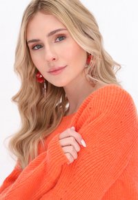 Pull en tricot orange vif avec une texture côtelée. Porté avec de grandes boucles d'oreilles florales rouges, montrant un gros plan du décolleté et de la main.