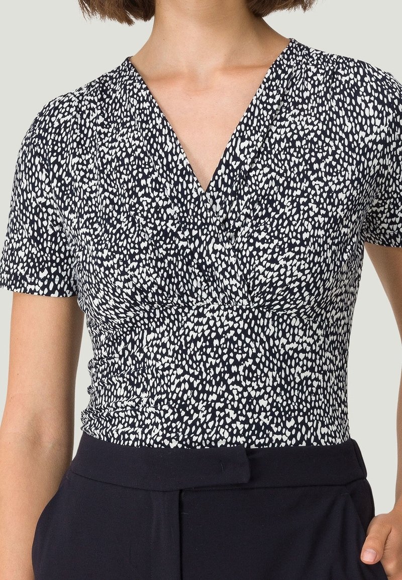 Haut bleu marine avec un motif à pois blancs, manches courtes, décolleté en V et détail froncé à la taille. Tissu lisse avec un léger éclat.