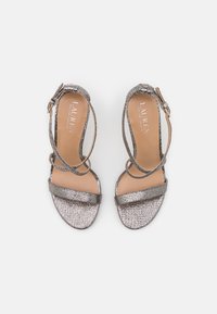 Metalliska silverhöga klacksandaler med korsade remmar, en bred framrem och spänne. Slät textur och vadderat fotbed.