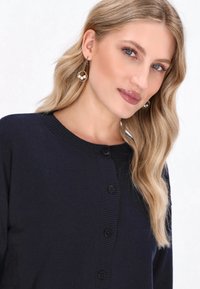 Námořnický cardigan vyrobený z měkké tkaniny s kulatým výstřihem, žebrovaným lemem a černými knoflíky na přední straně. Dlouhé, vlnité vlasy částečně viditelné.