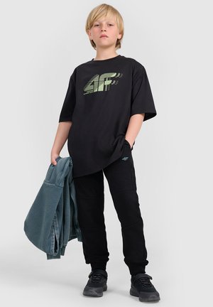 Blond dziecko stoi, nosząc czarną koszulkę z logo "4F", czarne spodnie, czarne trampki, trzymając w jednej ręce złożoną niebieską kurtkę.
