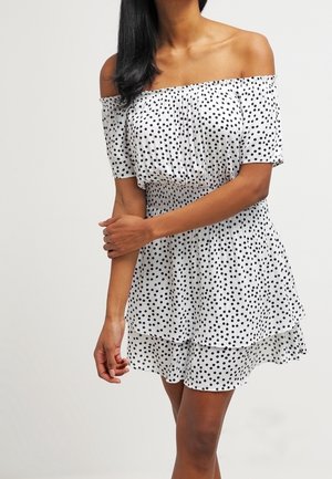 Femme portant une robe blanche épaules dénudées avec des pois noirs, les bras légèrement croisés, debout devant un fond uni.