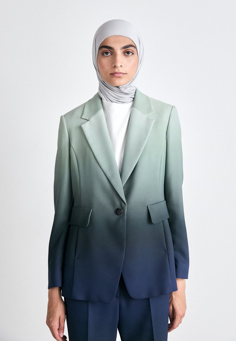 Blazer mit einem Farbverlauf von Grün zu Blau, mit Reverskragen, einreihiger Knopfschluss, zwei Vorderaschen, kombiniert mit einem grauen Hijab und einem weißen Hemd.