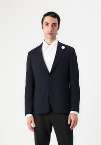 Mere tekstuuriga tumesinine bleiser, kahe nööbiga ja eespooltaskutega. Valge triiksärk, millel on bändkaelus ja väike valge lilleline pins.