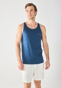 Canotta blu navy in morbido tessuto con scollatura rotonda e un piccolo logo dorato sul petto. Abbinata a pantaloni corti bianchi in lino.