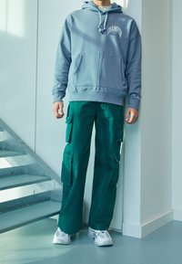 Šedá mikina s kapucí a přední kapsou, kombinovaná se zelenými cargo kalhotami s několika kapsami. Doplněno o bílé tenisky.