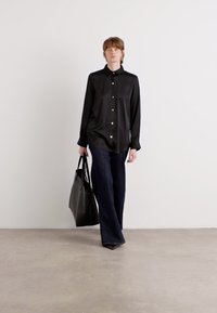 Camisa preta de botão com colarinho, combinada com jeans escuros de perna larga. O modelo leva uma bolsa tote preta e tem cabelo castanho.