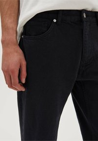 Pantalones vaqueros de mezclilla negra con un diseño clásico de cinco bolsillos, cinturilla resistente y detalles de costura visibles. El material parece texturizado y duradero.