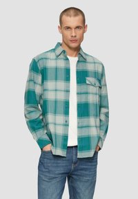 Zielona i szara koszula w kratę z długim rękawem, z kieszonką, połączona z białym t-shirtem i niebieskimi dżinsami.