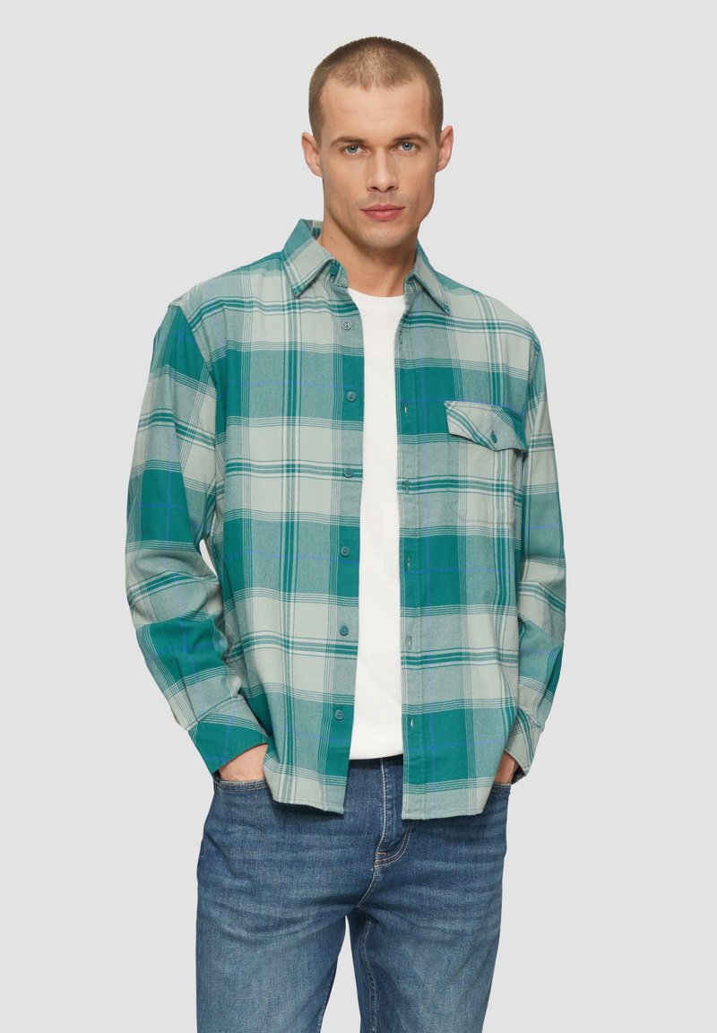 Zielona i szara koszula w kratę z długim rękawem, z kieszonką, połączona z białym t-shirtem i niebieskimi dżinsami.