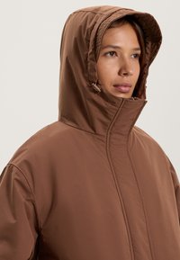 Bruine geïsoleerde jas met een capuchon, met een gladde, matte textuur en een hoge kraag. Zichtbare stikdetails benadrukken het ontwerp.