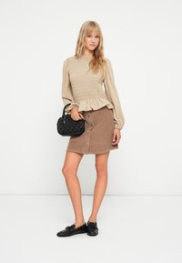 Blusa beige con fruncidos y mangas largas, combinada con una falda de pana marrón con botones. Sosteniendo un bolso negro texturizado. Zapatos negros.