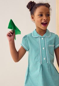 Grøn gingham-kjole med korte pufærmer, hvid kant og knapper. Barn holder et foldet grønt papirfly og smiler, mod en neutral baggrund.