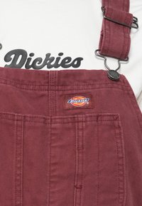 Salopette bordeaux en tissu durable, dotée de clips métalliques réglables, d'une poche avant et d'un patch logo brodé.