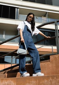 Salopette in denim blu, abbinata a una t-shirt bianca, elegante borsa grigia e scarpe platform bianche e spesse. La modella posa su scale all'aperto.