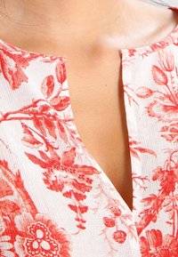 Blusa floreale con sfondo bianco e motivi floreali rossi, scollatura a V e tessuto testurizzato.